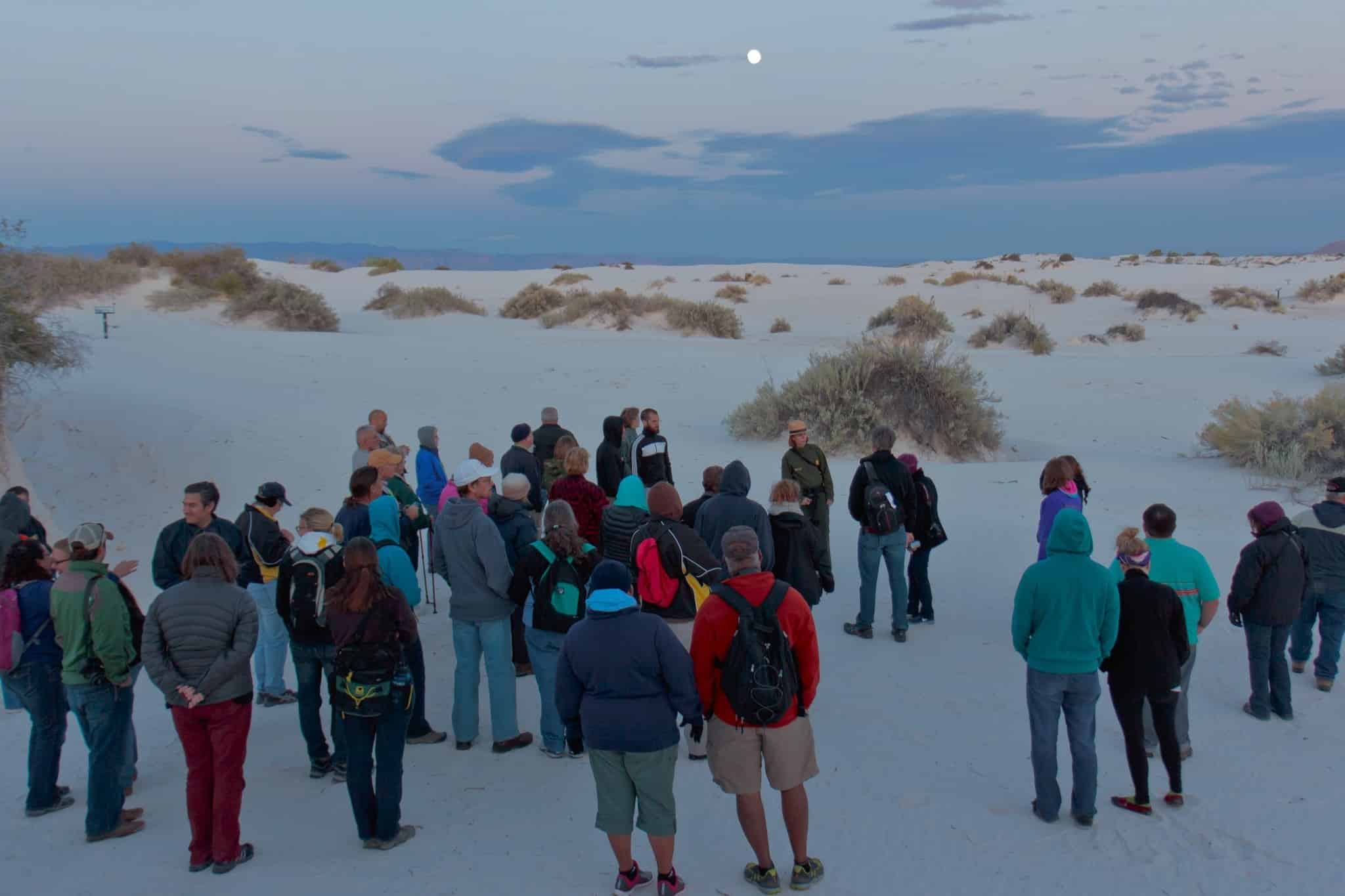 A group of people standing in the desert at dusk, attending one of the Night Sky Programs offered at White Sands National Park.